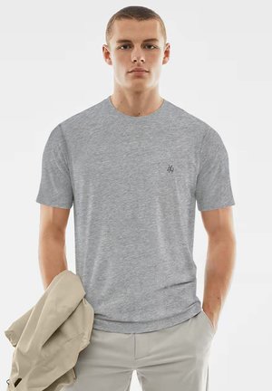 Graues T-Shirt mit kurzen Ärmeln aus weichem Stoff, Rundhalsausschnitt und kleinem schwarzem Logo auf der Brust, getragen von einem Modell, das eine Jacke hält.