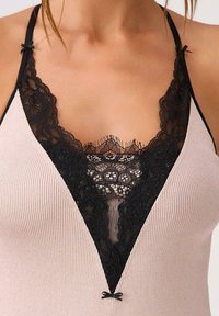 Camisole z koronkowym wykończeniem i głębokim dekoltem w kształcie litery V, wykonana z prążkowanej tkaniny w delikatnym różowym kolorze. Ozdobiona czarną koronką i małymi kokardkami.