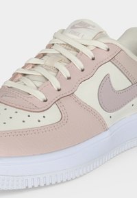 Κοντινή λήψη ενός ροζ και κρεμ Nike Air Force 1 αθλητικού παπουτσιού με λευκά κορδόνια και υφασμένο λευκό πέλμα.