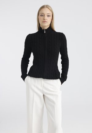 Femme portant un pull noir en maille torsadée zippé et un pantalon blanc à jambes larges, debout, les mains dans les poches, sur un fond uni.