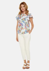 Blusa a stampa floreale con maniche a volant, tessuto leggero; abbinata a pantaloni bianchi aderenti e sandali bianchi con due cinturini.