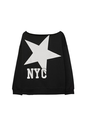 Vrouw die een zwarte off-shoulder top draagt met een witte ster en tekst "NYC", gecombineerd met zwarte uitlopende broeken en zwarte sneakers, staand tegen een bruine achtergrond.