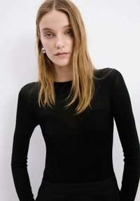 Joven con pelo largo y rubio lleva una camiseta negra ajustada de manga larga con un patrón texturizado, pendientes de aro plateados, de pie contra un fondo blanco.