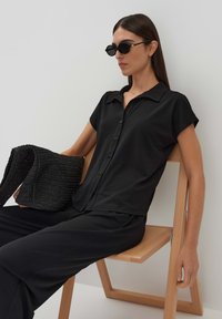 Zwarte kraagloze t-shirt met korte mouwen, vooraan knopen, gecombineerd met losse zwarte broeken en een zwart handtas met structuur.