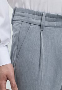 Gros plan d’une personne portant un pantalon plissé gris clair avec des poches latérales et un poignet de chemise blanche à manches longues visible près de la main.