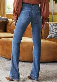 Jeans flare in denim azzurro chiaro con tasche posteriori, caratterizzati da una vita media e una finitura pulita all'orlo. Indossati con una blusa color ruggine.