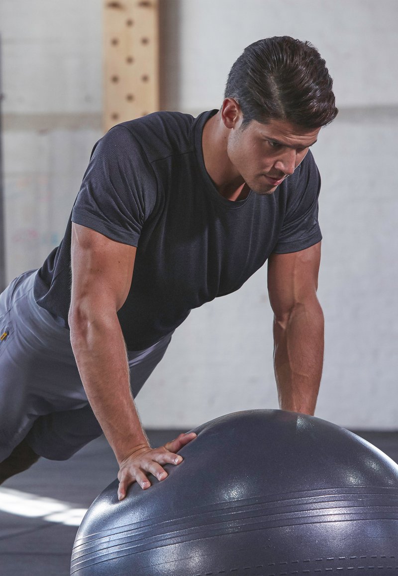Schwarzer Gymnastikball auf einem Fitnessboden, während ein Mann Liegestütze macht, gekleidet in einem enganliegenden schwarzen Shirt und grauen Shorts. Strukturierte Oberfläche sichtbar.