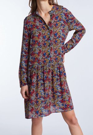 Frau trägt ein langärmliges, knielanges Kleid mit buntem Blumenmuster in Rot, Blau, Weiß und Senfgelb auf dunklem Hintergrund.