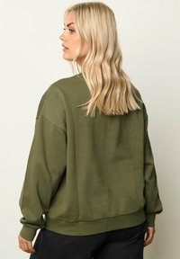 Maglione oversize verde oliva con scollo rotondo, polsini e orlo a costine, e un dettaglio grafico sul retro. Tessuto morbido con una texture opaca.