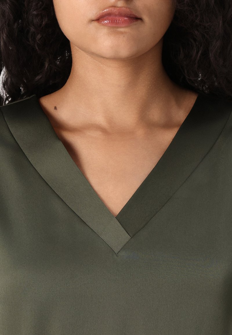 Camiseta verde oliva de cuello en V con una textura suave, que presenta un cuello ancho y un corte relajado. La tela parece ligera y ligeramente brillante.