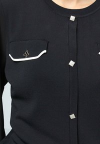 Haut en tricot noir à manches longues, doté de deux poches poitrine avec bord blanc, de détails en strass carrés et d'une fermeture à boutons sur le devant.