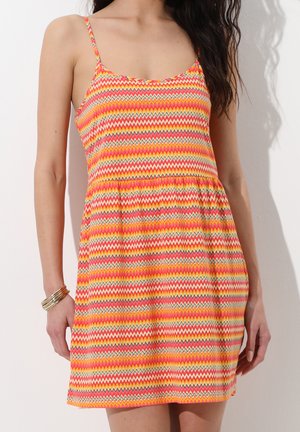 Donna con un vestito senza maniche dal motivo zigzag colorato, con spalline sottili e una vita ampia e arricciata, in piedi contro un muro semplice.