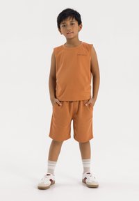 Pomarańczowa bezrękawnikowa koszulka z małym detalem tekstowym, w zestawie z dopasowanymi szortami; obie części mają miękką teksturę. Białe sneakersy z czerwonymi akcentami.