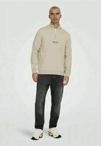Uomo con capelli biondi corti che indossa un dolcevita beige a zip, jeans neri e sneakers bianche, in piedi su uno sfondo neutro.