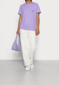 T-shirt in cotone lavanda con maniche corte e un logo sul petto, abbinato a pantaloni a gamba ampia color crema e una borsa texture lavanda. Sneakers bianche.