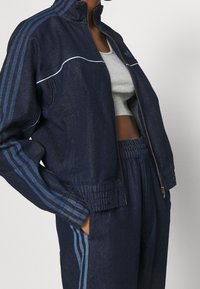 Person i en marinblå Adidas-träningsdräkt med ljusblå ränder och en grå cropped tanktop, händerna i fickorna, jackan halvdraget.