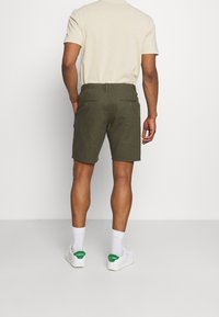 Pantaloni corti verde oliva realizzati in tessuto leggero. Vita elasticizzata con coulisse e due tasche posteriori. Indossati con sneaker bianche e calzini.