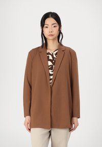 Hnedý blazer s uvoľneným strihom, klopovými goliermi a dlhými rukávmi, nosený cez vzorovaný top; hladká látka a minimálne detaily.