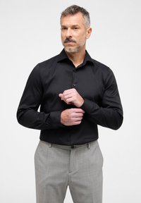 Camicetta nera a maniche lunghe con colletto a bottone e tessuto liscio, abbinata a pantaloni grigi chiari con texture.