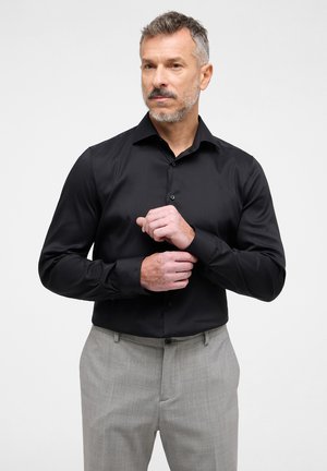 Schwarz langärmliges Hemd mit Button-Down-Kragen und glatter Textur, kombiniert mit hellgrauen strukturierten Hosen.