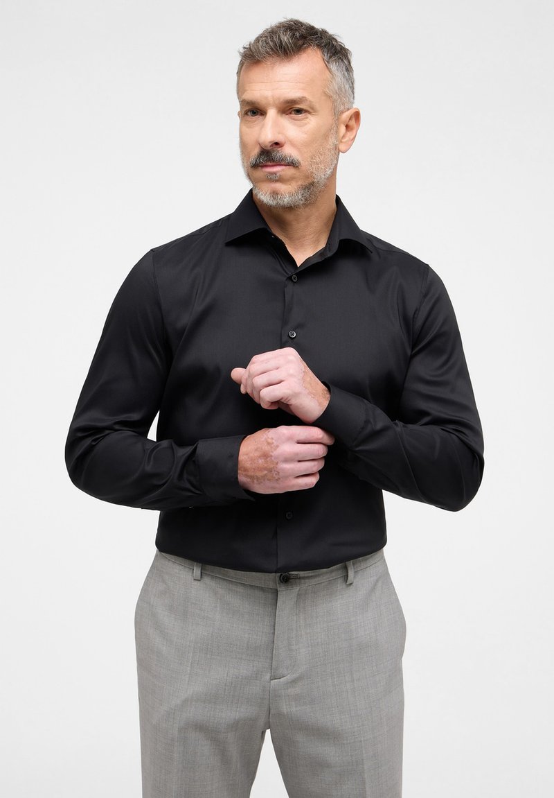 Camicetta nera a maniche lunghe con colletto a bottone e tessuto liscio, abbinata a pantaloni grigi chiari con texture.
