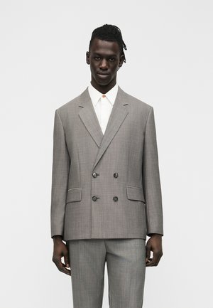 Muž s tmavou pokožkou a vrkočmi, oblečený v svetlosivom dvojradovom obleku a bielej košeli, stojaci pred jednoduchým bielym pozadím.