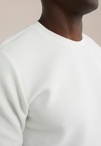 Vit texturerad sweatshirt med ribbad rund halsringning och en närbild på tygets vävda mönster och släta yta.