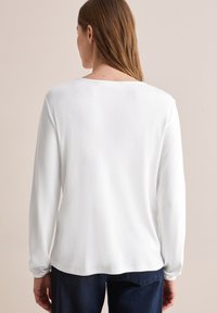 Witte lange mouwen top met een ronde halslijn en geplooide manchetten, gemaakt van zachte stof. Draag het met donkerblauwe broek, met minimale naaddetails.