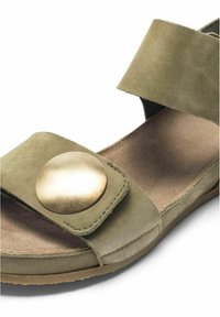 Sandale en daim vert olive avec de larges brides, dont une avec un grand bouton métallique rond, et une semelle plate coussinée.