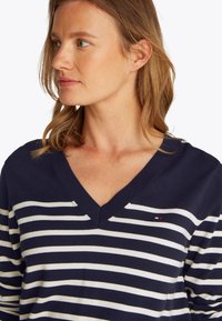 Kobieta o jasnobrązowych włosach, ubrana w granatowo-biały sweter w paski z dekoltem w serek, patrząca w lewo na jednolitym białym tle.