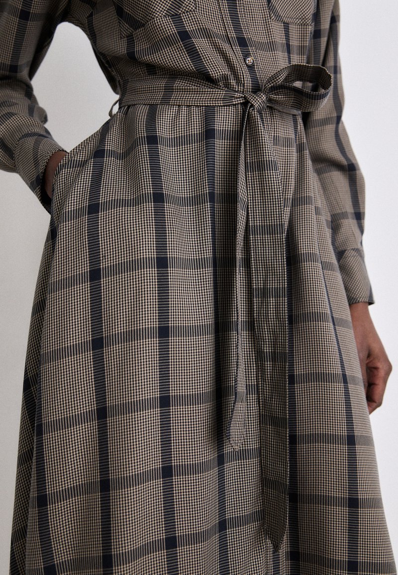 Langes kariertes Kleid in Beige und Marineblau, mit langen Ärmeln, einem Bindegürtel in der Taille und Taschen, das ein schachbrettartiges Muster aufweist.