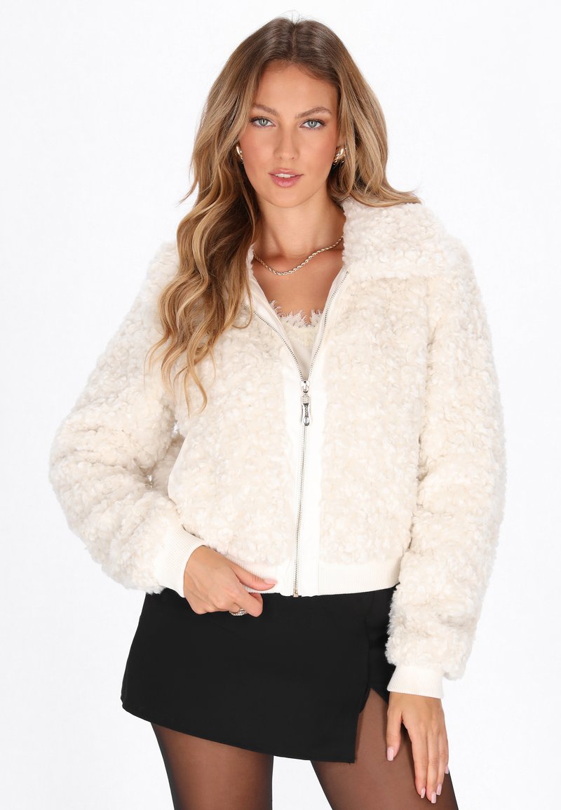 Femme aux longs cheveux blonds portant une veste zippée texturée crème, une mini-jupe noire, des collants noirs et un collier en chaîne dorée sur fond blanc.