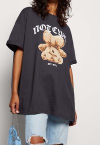 T-shirt oversize grigio scuro con una grafiche di un orsetto cartoon. Presenta il testo in grassetto "NON CUTE MA SELVAGGIO" sopra e sotto l'immagine. Materiale in cotone.