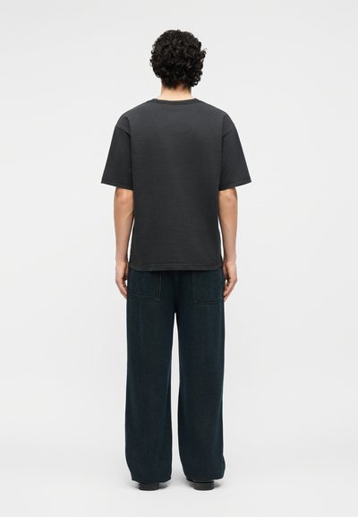 Άτομο με σγουρά μαλλιά στέκεται με την πλάτη, φοράει απλό μαύρο κοντομάνικο πουκάμισο και σκούρο παντελόνι με φαρδιά πόδια και πίσω τσέπες.