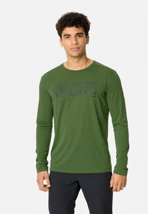 Giovane uomo con capelli corti e ricci indossa una maglia verde a maniche lunghe con un motivo grafico e pantaloni scuri, in piedi davanti a uno sfondo semplice.