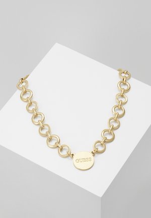 Collier chaîne en or avec de gros maillons circulaires et un pendentif rond gravé du mot "GUESS" sur un bloc d'exposition blanc.