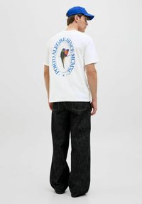 Camiseta blanca de manga corta con un gráfico colorido de loro en la espalda, combinada con jeans de denim oscuros de corte holgado y una gorra azul.