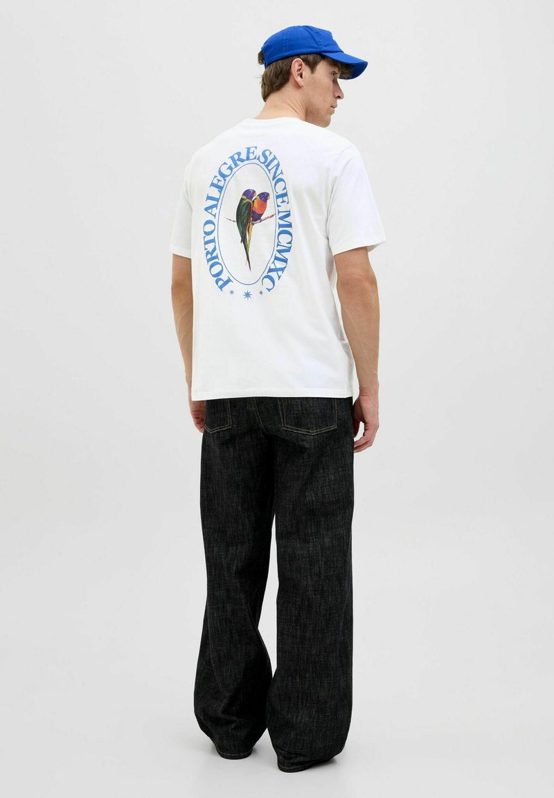 Camiseta blanca de manga corta con un gráfico colorido de loro en la espalda, combinada con jeans de denim oscuros de corte holgado y una gorra azul.