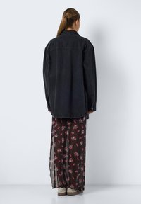 Överdimensionerad svart denimjacka över en genomskinlig svart blommig maxiklänning; klänningen har röda och gröna blommönster, med en mjuk textur och flödande silhuett.