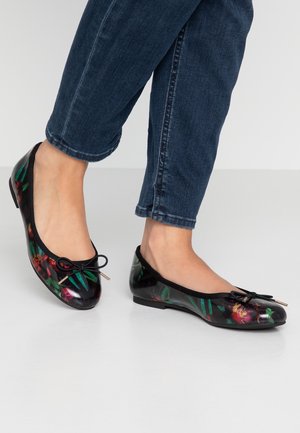Pieds portant des ballerines à motif floral foncé associées à un jean en denim bleu foncé retroussé, debout sur un sol blanc.