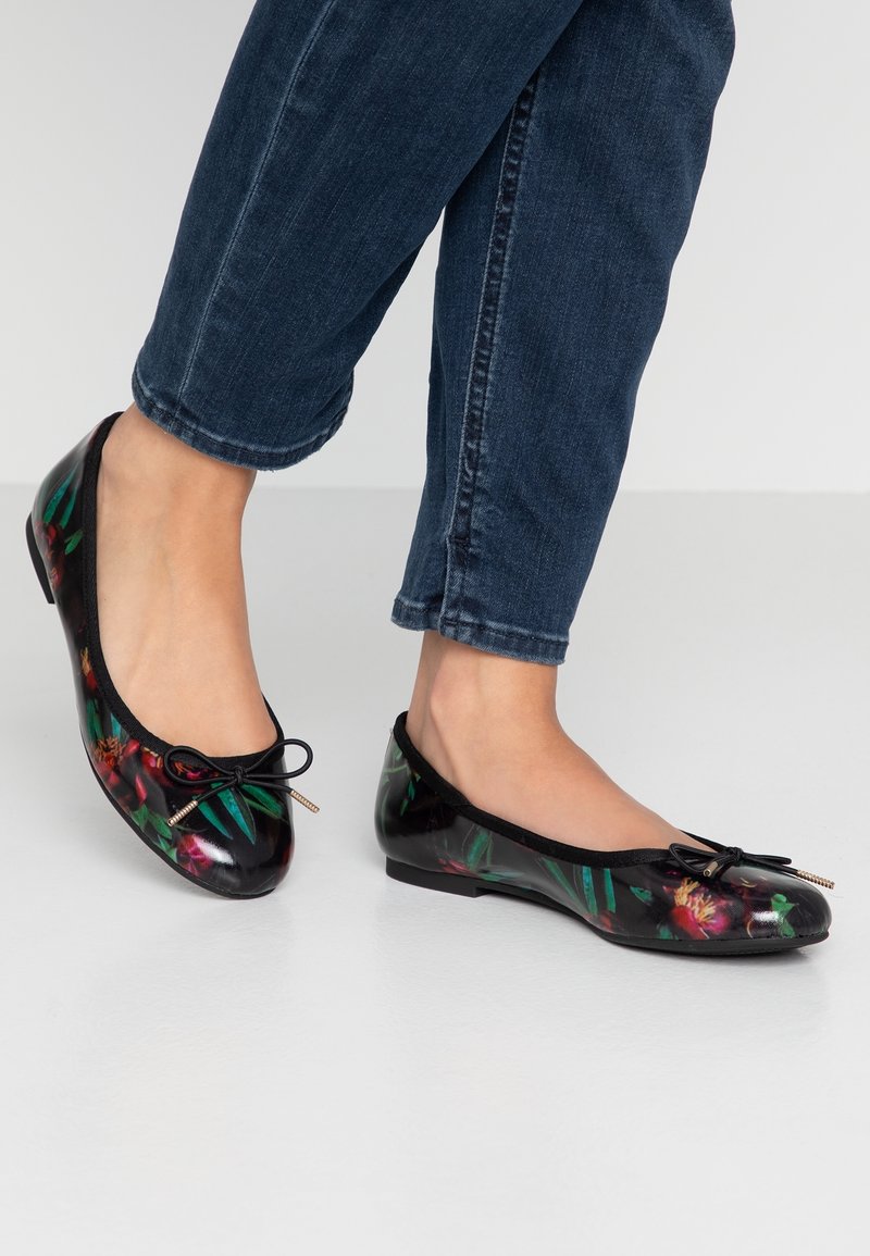 Pieds portant des ballerines à motif floral foncé associées à un jean en denim bleu foncé retroussé, debout sur un sol blanc.