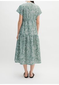 Femme portant une robe midi verte à motif floral blanc et manches courtes, debout de dos, portant des sandales plates noires.