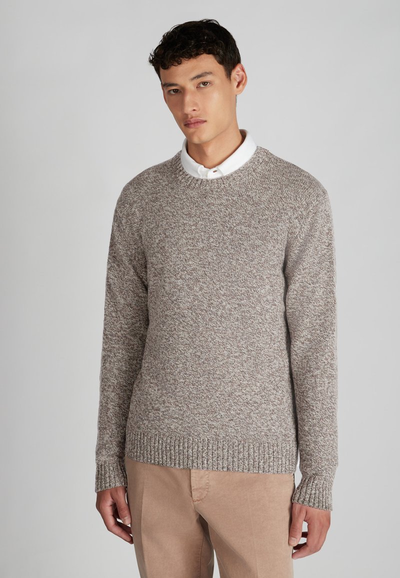 Pull en maille marron avec un col rond et des poignets côtelés, porté sur une chemise blanche à col, associé à un pantalon beige clair.