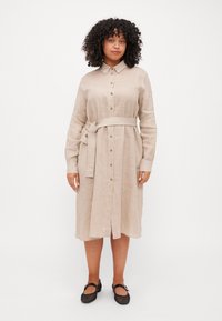 Abito camicetta in lino beige con colletto, bottoni frontali, maniche lunghe e una cintura di stoffa. Caratterizzato da un taglio al ginocchio e sottili spacchi laterali.