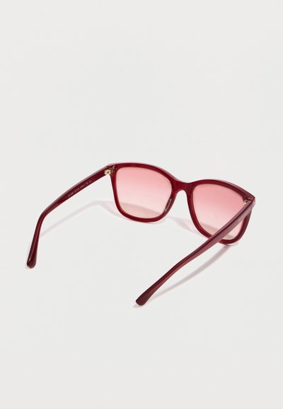 Γυαλιά ηλίου με δέσμη σε σχήμα γάτας, με γυαλιστερούς κόκκινους σκελετούς και ροζ φακούς με διαβαθμισμένο χρώμα, ανοιχτά πάνω σε λευκή επιφάνεια.