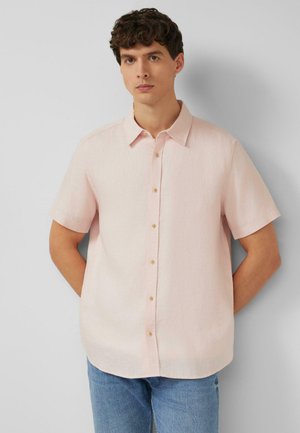 Młody mężczyzna z krótkimi kręconymi włosami, ubrany w jasnoróżową koszulę z krótkim rękawem na guziki i niebieskie dżinsy, stoi na tle jednolitego szarego tła.