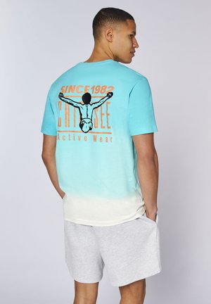 Man wearing blue and white gradient T-shirt with orange and black graphic and gray shorts, standing sideways with hands in pockets.