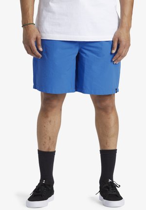 Homme portant un short bleu vif, des chaussettes noires arrivant aux mollets et des baskets noires, se tenant devant un fond blanc.