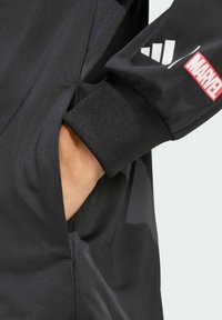 Black long-sleeve garment with a pocket and ribbed cuffs; features white and red logos from Adidas and Marvel. Smooth texture, athletic design.