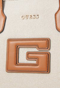 Lærredstaske med beige struktur, prydet med et guld "GUESS"-logo og en brun læderdetalje formet som et G. Ren design med kontrasterende farver.